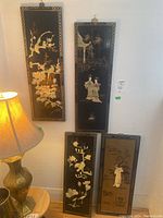 Four lacquered Asian decorative panels shown vertically on a wall next to a lamp, displaying varied traditional scenes with birds, flowers, and figures.