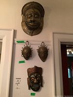 Group shot of four masks mounted on wall: large brass mask at top, two small sunburst metal masks in middle, carved wood/copper mask at bottom