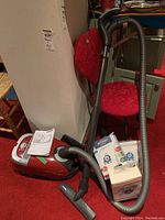 Red Miele canister vacuum with attached hose and wand, floor tool on red carpet, box containing filters, manual on top
