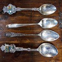 Top view of four sterling silver collectible spoons laid out on wooden surface showing variety of designs, enamel crests, and engravings on bowls.