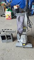 Kirby vacuum cleaner with cloth dust bag, attached power cord, and separate attachment pieces on a concrete floor with other items in background.