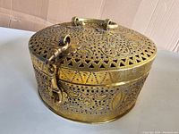 Exterior of round brass jewelry box with hinged lid, handle, latch, and pierced Jali cut floral pattern