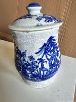 Side view of the blue and white ceramic jar with lid showing traditional Chinese figure and landscape design, crackle glaze visible.