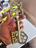 Photo of an extra-large vintage brass wall key with ornate scroll details in the bow and clover-shaped opening being held