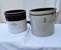 Pair of antique stoneware crocks, one with a crown marking and one marked '3' showing minor crack, displayed on white fabric.