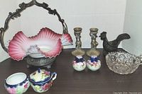 Front view of antique bridal basket with pink and white ruffled glass, two broken pieces visible, vintage pewter candlesticks, crystal candy dish, salt and pepper shakers, and cast iron rooster ashtray on surface