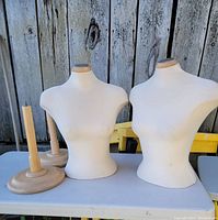 Two mannequin torsos along with two wooden pedestals on a table against a rustic wooden fence background