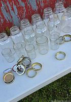 Twelve mason jars arranged on white surface with various gold-colored metal lids and rings next to them.