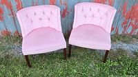 Two pink side chairs placed on grass outdoors, showing front views with tufted backs and wooden legs.