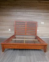 Front view of the assembled queen bed frame showing cherry wood slat headboard and solid footboard on a grey floor with wood panel wall behind.