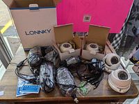 Photo showing two wired dome security cameras out of the box with cables, power adapters, mounting accessories, and product literature on a table.