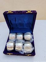 Photo showing six silver napkin rings inside a purple velvet hinged box with blue interior lining. Rings wrapped in plastic.