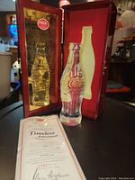 Crystal Coca-Cola bottle displayed in open wooden box with certificate of authenticity placed nearby.