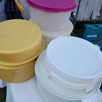 Stack of vintage Tupperware containers showing yellow, white, and cream colors with assorted lids including purple. Containers are round and vary in size.