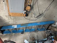 Blue aluminum Werner step ladder, 8 feet long, folded and laid horizontally on a concrete basement floor near electrical panel and miscellaneous items.