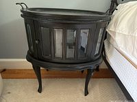 Black oval wooden nightstand with beveled edge tray top, glass inset front door panels, curved decorative legs, and metal handles on each side.