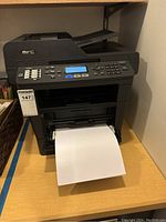Front view of Brother MFC multi-function printer with paper output tray showing paper printed out. Device control panel visible with display and control buttons.