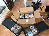 Photo showing two black zippered DVD storage cases open with DVDs inside and a cardboard box containing DVD cases on a table