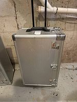 Front view of a silver aluminum craft suitcase standing upright, showing the front panel and a black handle on top.