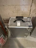 Closed silver aluminum crafts case with black handle and metal latches, standing on concrete floor against a block wall.
