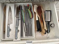 Drawer containing three Misen brand kitchen knives with gray handles in boxes, multicolor wire whisk, metal wire whisk, vegetable peeler with green handle, wooden and plastic spatulas, metal grater with black handle, and metal scoop.