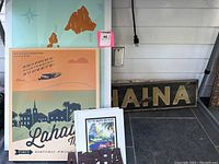 Photo showing Hawaiian travel posters including a map and a vintage style Lahaina Maui poster with retro colors, plus a small framed travel print and a wooden Lahaina sign leaning against a wall.