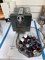 Front and side angle showing the espresso machine, wire basket full of assorted colored espresso pods, and the Nespresso manual.
