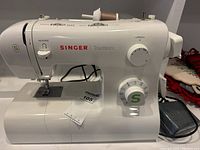 Front side view of white Singer Tradition sewing machine showing stitch length and stitch pattern dial, reverse lever, and spool holder with brown thread.