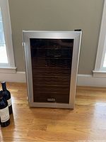 Danby wine fridge with silver framed glass front door closed, standing on hardwood floor with some wine bottles next to it (not included).