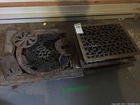 Four brass rectangular forced air floor grates with patterned openwork design and some additional metal parts visible on the left grate.