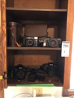 Photo shows two shelves inside a wooden cabinet containing cameras, binoculars, and cases.