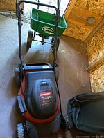 Red and black Troy-Bilt 48V cordless lawnmower with black grass bag attached, seen from front angle inside a wooden shed.