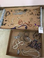 Top view showing an assortment of costume jewelry including necklaces, bracelets, and many small pairs of earrings scattered and organized loosely in two cardboard boxes.