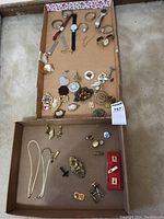 Full overview of all items including watches, brooches, tie clips, and earrings in two cardboard boxes arranged on carpet.
