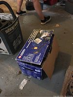 Packaged Campbell Hausfeld pressure washer box in a garage setting with legs of a person passing by; box is closed but not sealed, indicating the contents are inside potentially.
