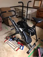 Full view of Vitamaster Fitness Bike, DP Gravity Body Lift machine, and weights including dumbbells and exercise handles on beige carpet floor with furniture in background.
