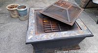 Outdoor square metal firepit with decorative tiled border, mesh cover resting on top and grill grate inside