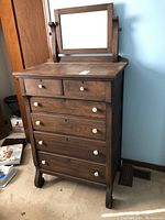 Front view of tall wooden highboy dresser with mirror, showing six drawers and white round knobs.