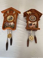 Two wooden limited edition cuckoo clocks; one Thomas Kinkade Timeless Memories with cottage art, and one Pugs Make A Home A Home themed clock with pug images and dog figure on roof.