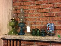 Front view showing two oil lamps with chimneys, extra chimney, insulators and lighthouse candle holder on brick surface.