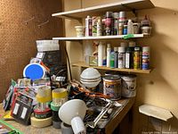 Wide view of paint cans, tapes, lamps, cords and switch covers displayed on a workbench and shelves.