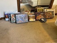 Wide view of collection of assorted puzzle boxes arranged on carpeted floor underneath a mirror. Puzzles are from various brands and show a range of artwork including cityscapes and animals.