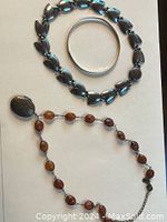 Three pieces: a sterling silver necklace with heart-shaped links, a sterling silver bangle, and a necklace with amber stones and locket pendant laid flat on a white surface.