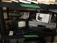 Overview of all electronics including VHS tapes, Curtis black and white TV, Durabrand portable DVD player with box, and the Signature and Citizen VHS players on a black shelf.