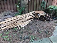 Wide view showing stacked mixed wood pieces of various lengths and thicknesses leaning on a fence near a tree.