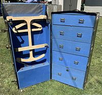 Open view of antique metal wardrobe steamer trunk showing blue fabric-lined interior with wooden clothes hangers and adjustable bars on left side and multiple drawers on right side.