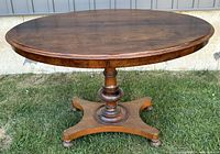 Front view of antique oval solid wood table on grass showing turned central pedestal and splayed foot base