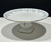 Front-side view of the vintage Royal Albert Memory Lane pedestal cake stand showing blue floral decoration and gold trim on the wide flat top and pedestal base.
