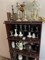 Full view of the shelving unit with all items displayed: metal candlesticks on top shelf, white hobnail glass candlesticks on middle shelf, decorative vessels and lamp on bottom shelves.