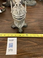 Front view of miniature knight's helmet armour on black stand with measuring tape showing height about 7 inches, condition visible with detailed embossed design